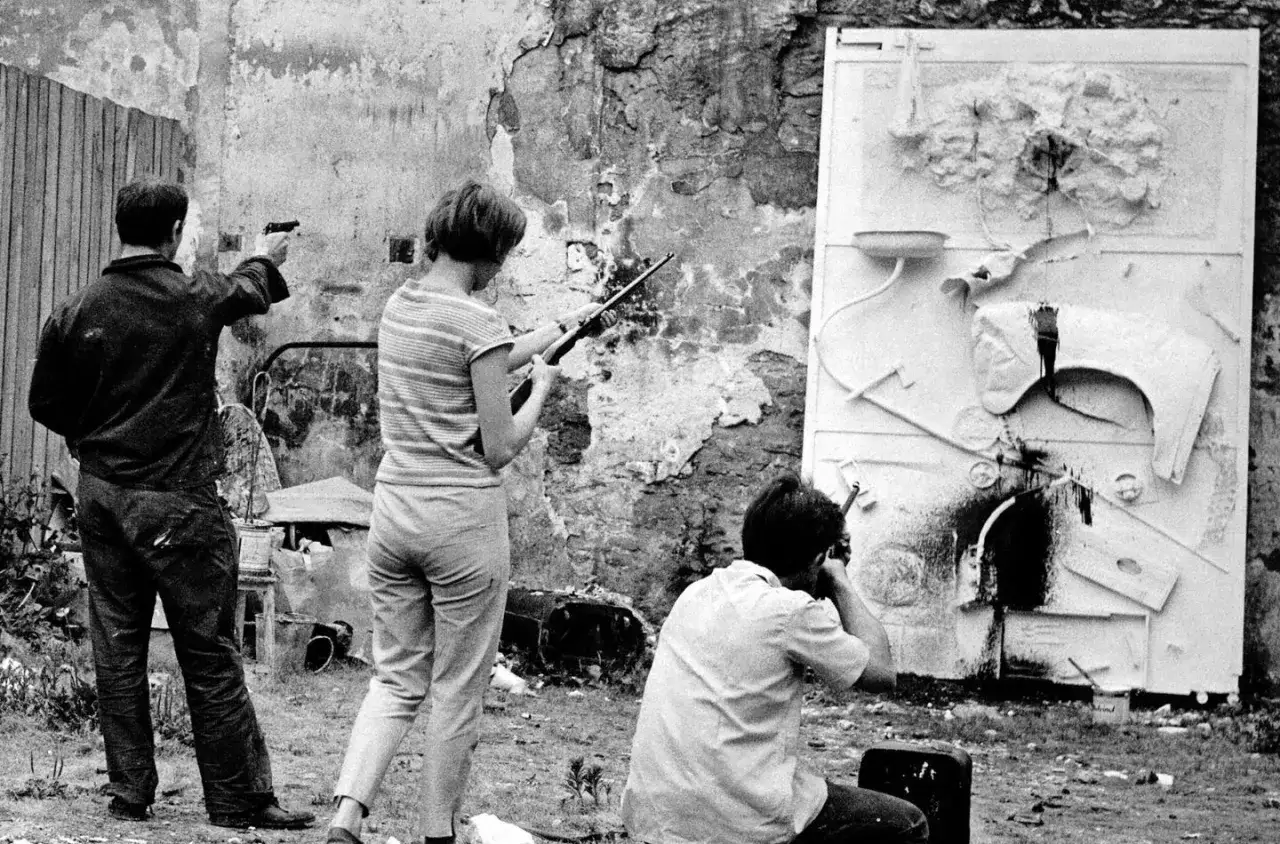 Trois personnes effectuent des tirs sur une sculpture. Un homme vise avec un pistolet, une femme avec une carabine, et un autre homme est accroupi, tirant sur l'&oelig;uvre de Niki de Saint Phalle.