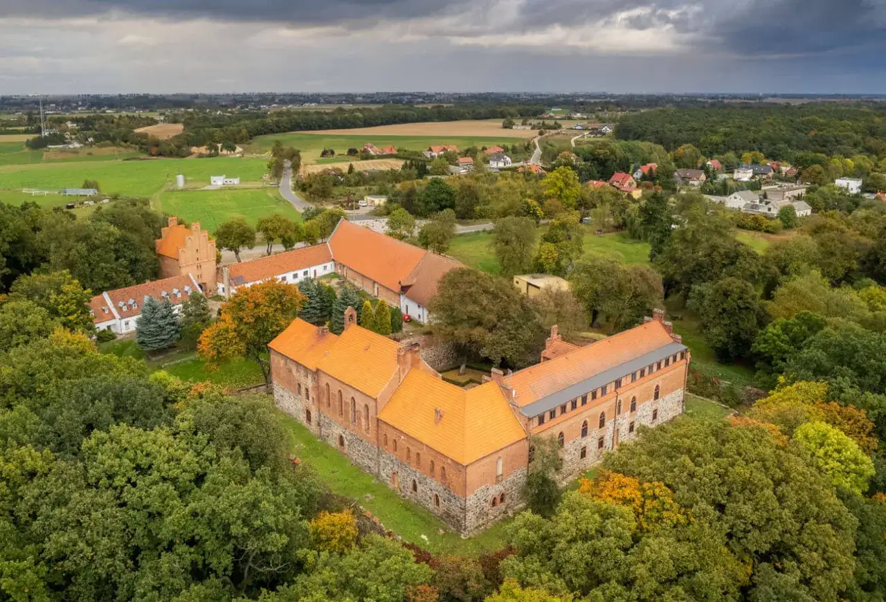 Zamek Bierzgłowski otoczony jesienną zielenią i polami. Ceglane mury i pomarańczowe dachy tworzą malowniczy krajobraz.