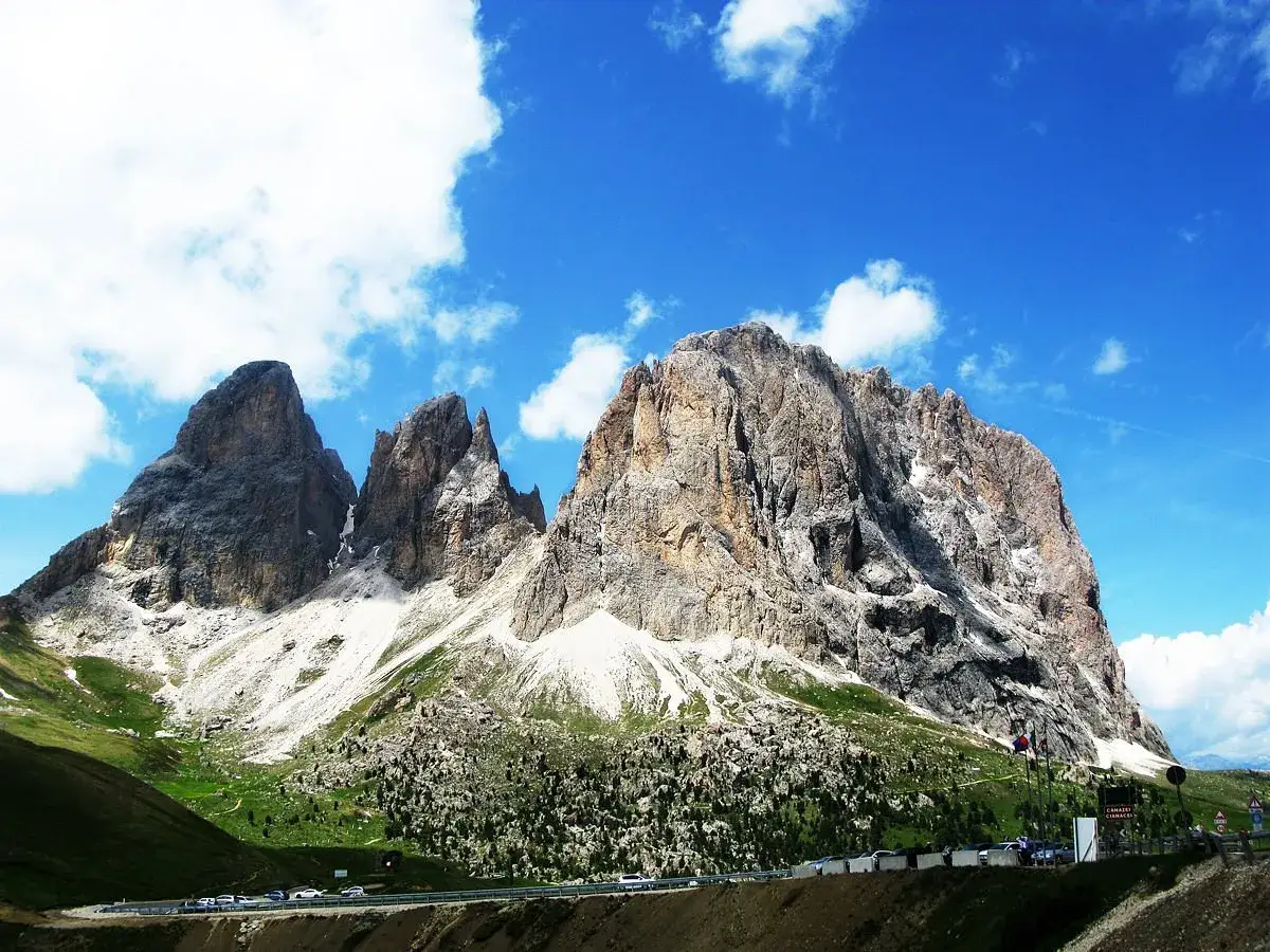 Imponenti vette rocciose del **Passo Gardena** sotto un cielo azzurro con nuvole bianche. Un nastro d'asfalto serpeggia ai piedi delle montagne.