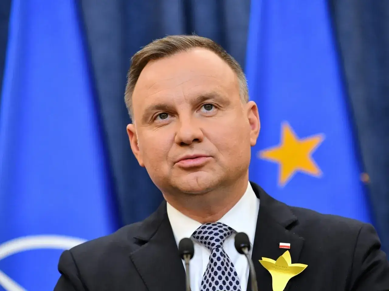 Prezydent Andrzej Duda, którego wiek nie jest widoczny na zdjęciu, przemawia na tle flag UE i NATO.