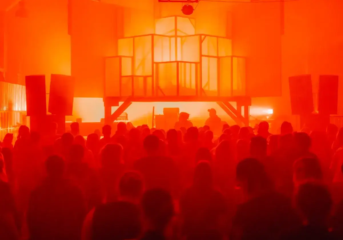 Tłum ludzi na rave festivalu, otoczony pomarańczowym światłem. Na scenie widać konstrukcję i sylwetki DJ-&oacute;w.