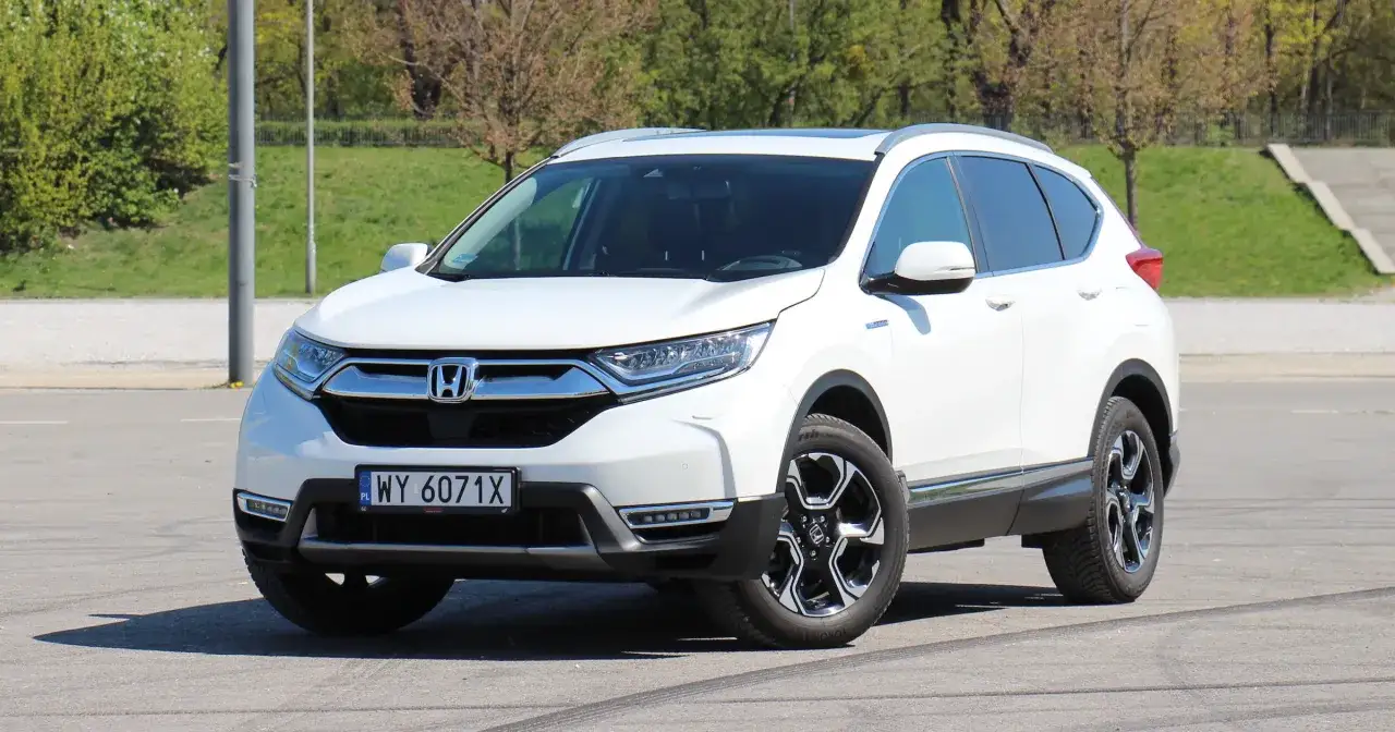 Biała Honda CR-V Hybrid zaparkowana na zewnątrz, z zielonym tłem i drzewami.