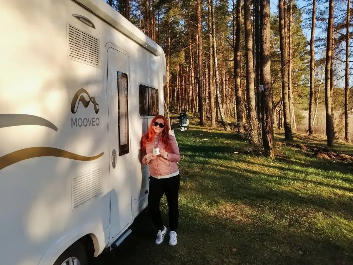 Kobieta z rudymi włosami i kubkiem kawy przy kamperze w lesie. Ciekawe miejsca na Ukrainie czekają na odkrycie!
