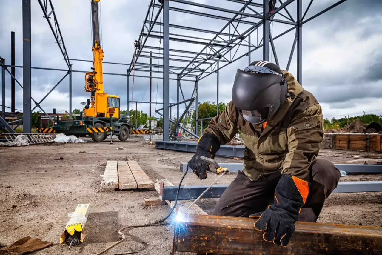 Operaio salda travi metalliche in cantiere, un lavoro duro che molti rifiutano. Gru gialla sullo sfondo.