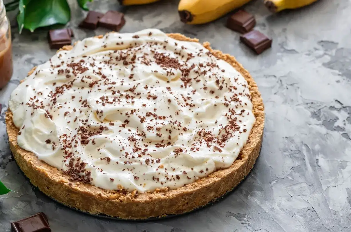 Puszysty banoffee pie z czekoladową posypką, obok banany i kawałki czekolady. Idealny deser, gdy masz ochotę na coś słodkiego, a nie na tartę cytrynową.