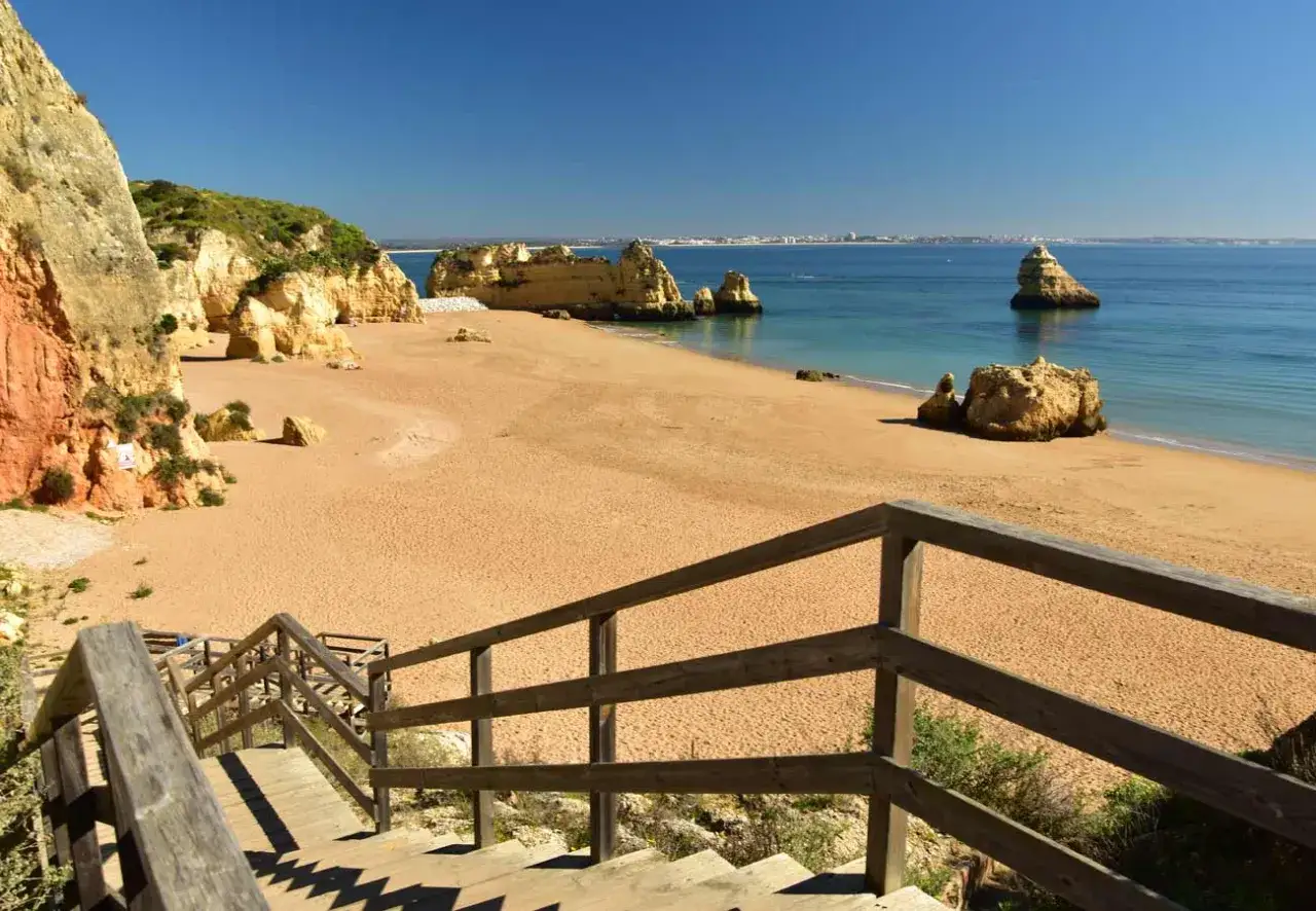 Holztreppe führt zu einem Sandstrand mit Felsformationen und Blick auf die Stadt Lagos am Horizont.