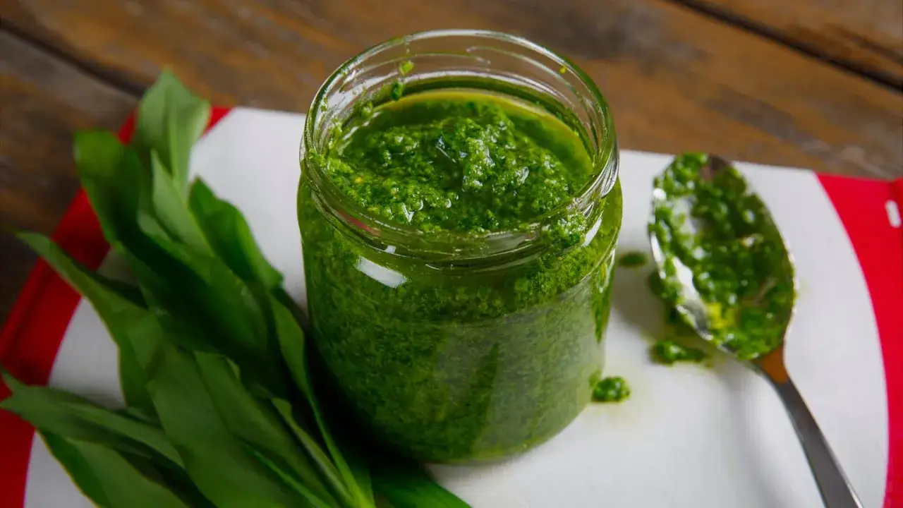 Słoik pełen aromatycznego pesto z czosnku niedźwiedziego, obok garść świeżych liści i łyżeczka sosu.