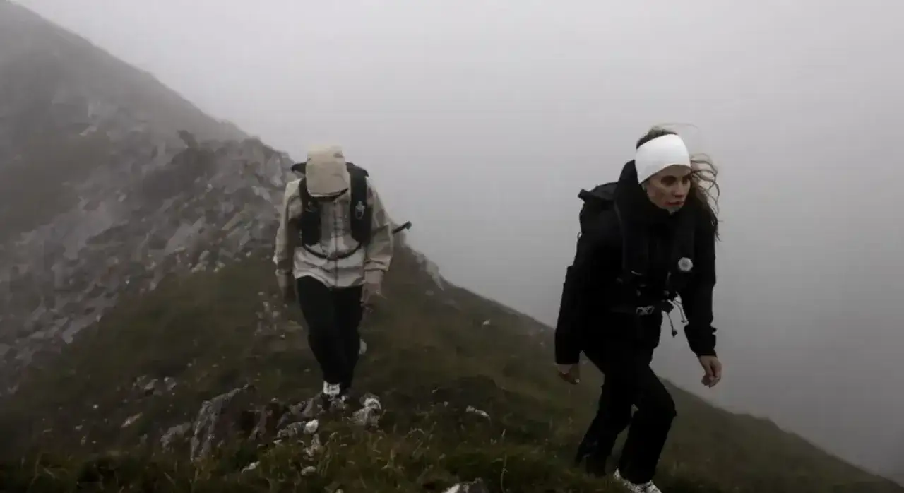 Dwie osoby wspinają się po zboczu góry w gęstej mgle. W oddali widać skaliste szczyty, a pogoda zapowiada burzę w górach.