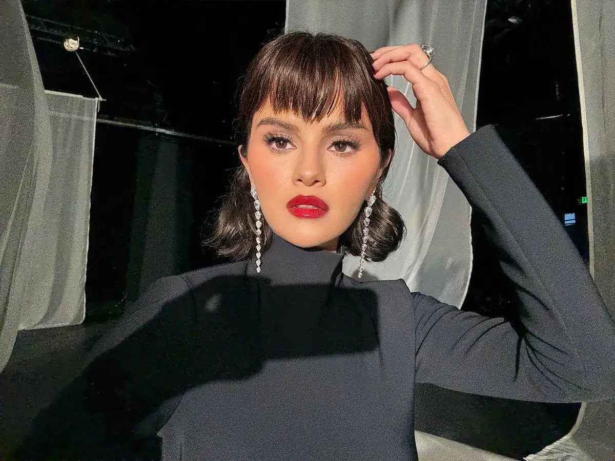 Donna con un elegante abito nero e una **frangia corta** che incornicia il viso, illuminato da un rossetto rosso acceso e orecchini pendenti scintillanti.