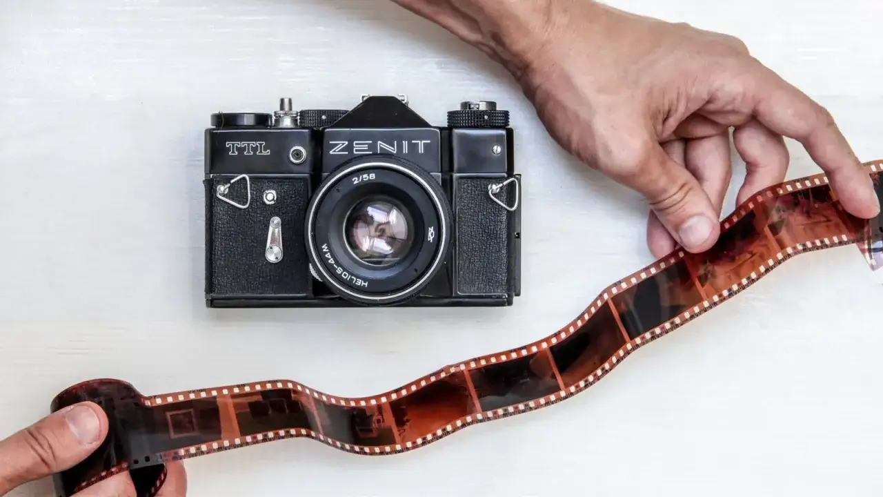 Dwie dłonie trzymają kliszę filmową obok aparatu Zenit TTL. Powrót do korzeni fotografii analogowej.