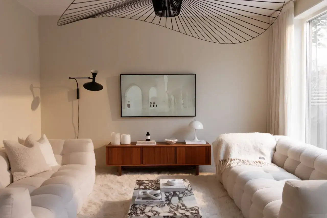 Ett modernt vardagsrum med vita soffor, en marmorb&auml;nk och en stor taklampa. Rummet pr&auml;glas av ljusa toner och en lugn atmosf&auml;r.