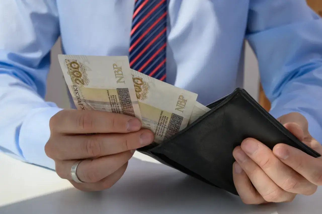 Mężczyzna w koszuli i krawacie wkłada do portfela banknoty 200 zł. To może być jego odprawa pośmiertna, symboliczne pożegnanie z pracą.