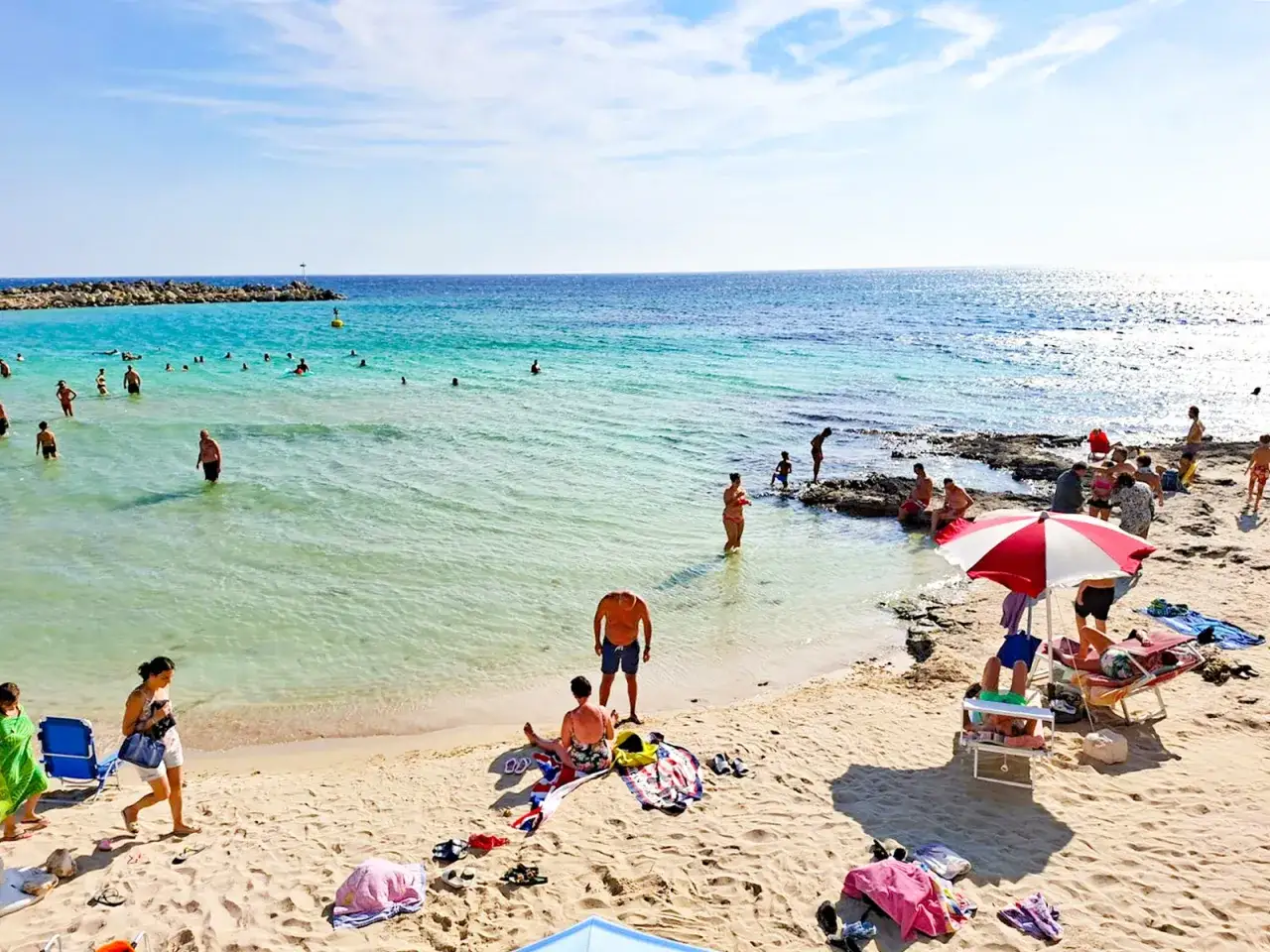 Spiaggia affollata con persone che nuotano e prendono il sole. In lontananza, una diga di rocce e il mare azzurro. La **torre vado centro storico** &egrave; un luogo ideale per godersi l'estate.