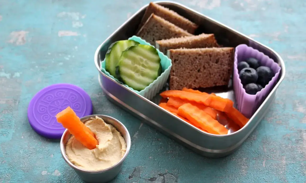 Gesunde und ungesunde Lebensmittel f&uuml;r den Kindergarten: Vollkornbrot, Gurkenscheiben, Karottensticks mit Hummus, Blaubeeren.