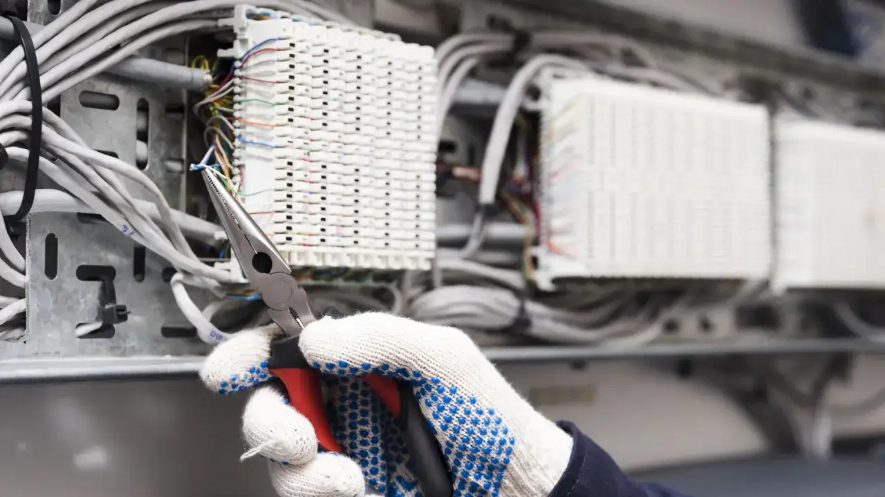 Ręka w rękawicy z kombinerkami pracuje przy instalacji niskoprądowej, łącząc przewody.
