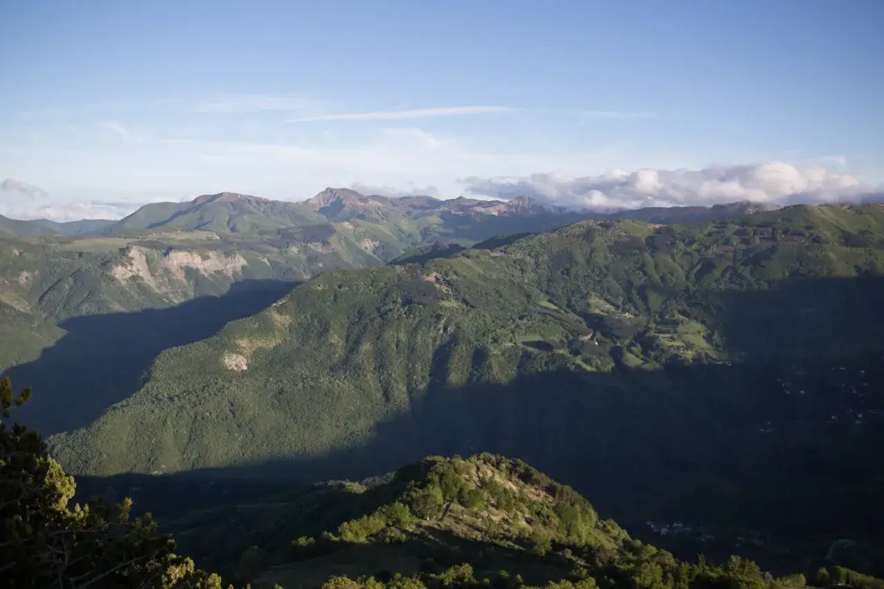 Vaste montagne verdi sotto un cielo azzurro, con nuvole che sfiorano le cime. Si intravede il profilo del monte più alto dell'Emilia Romagna.