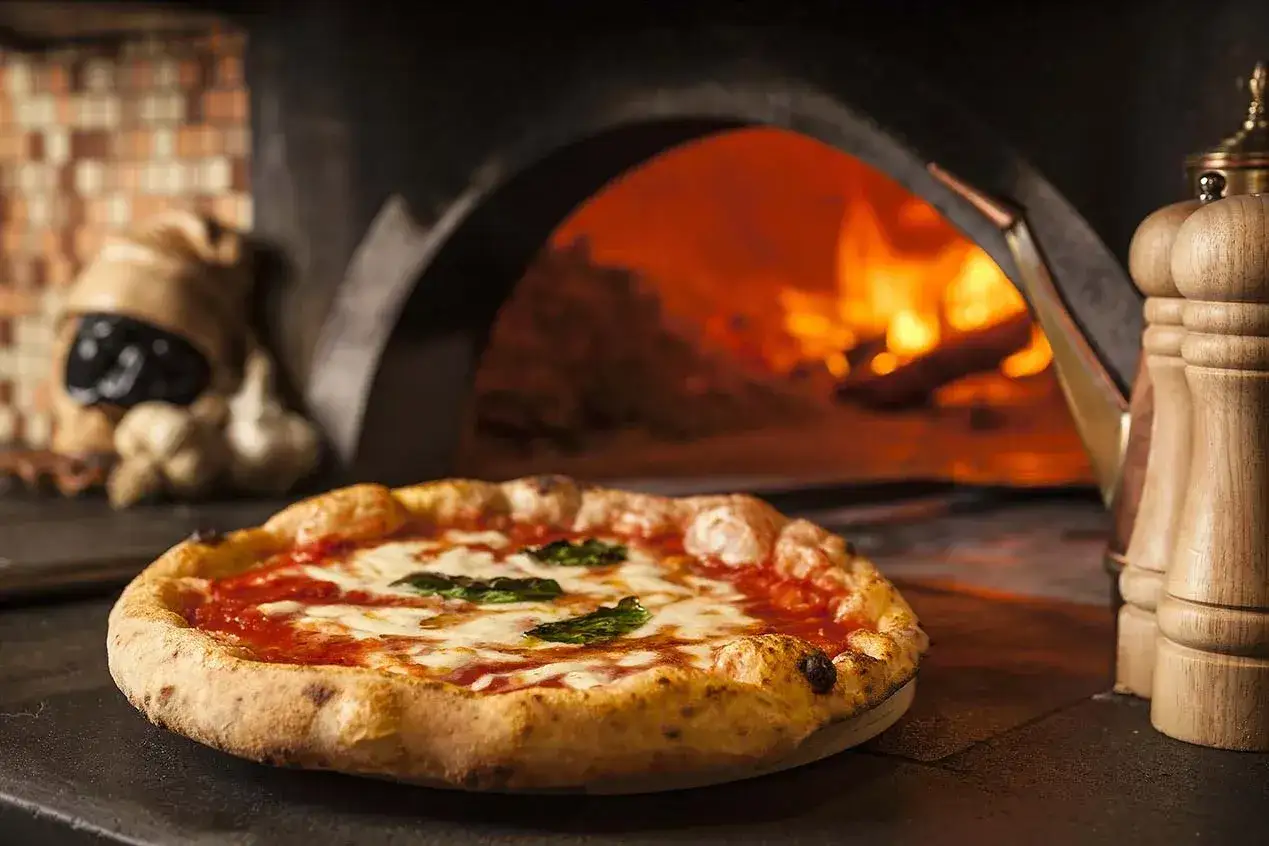 Pyszna pizza Margherita z pieca opalanego drewnem. W tle żar ognia i tradycyjne rodzaje pizzy.
