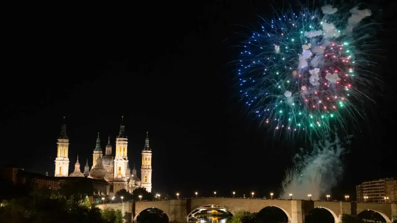 Espectaculares fuegos artificiales iluminan el cielo nocturno sobre el río Ebro, celebrando las fiestas del Pilar. La Basílica del Pilar se alza majestuosa, reflejándose en el agua.