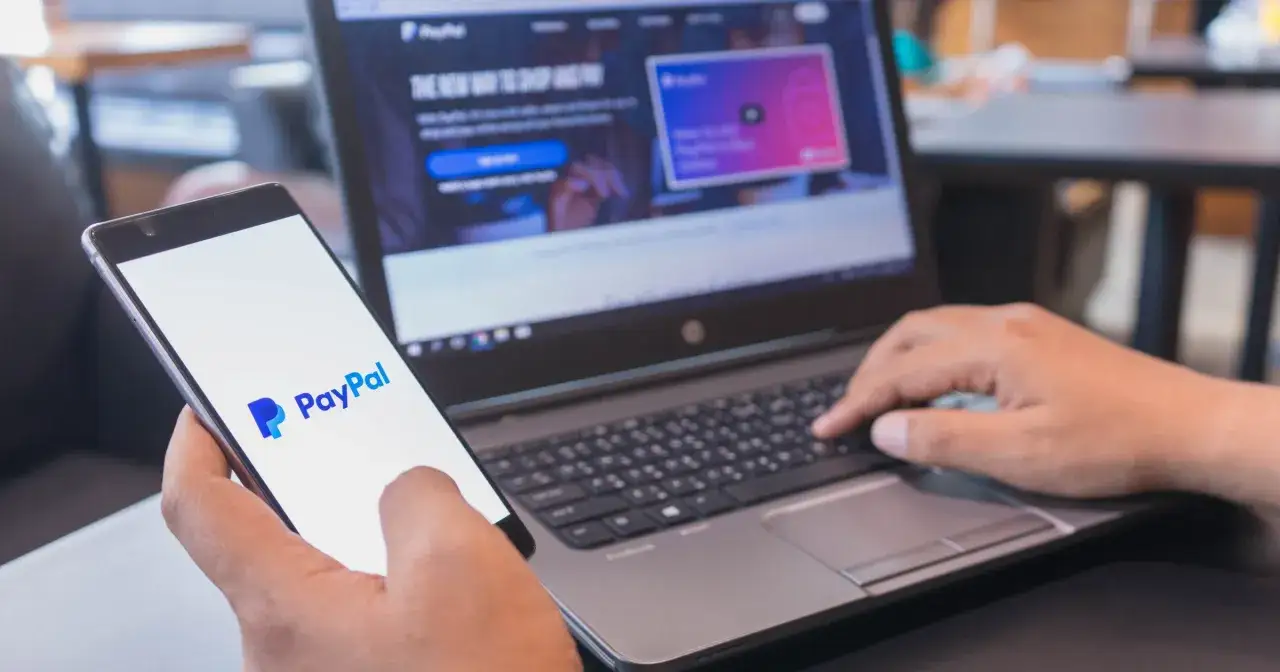 Osoba trzyma telefon z logo PayPal, a obok klawiatura laptopa. Dowiedz się, paypal co to i jak działa.
