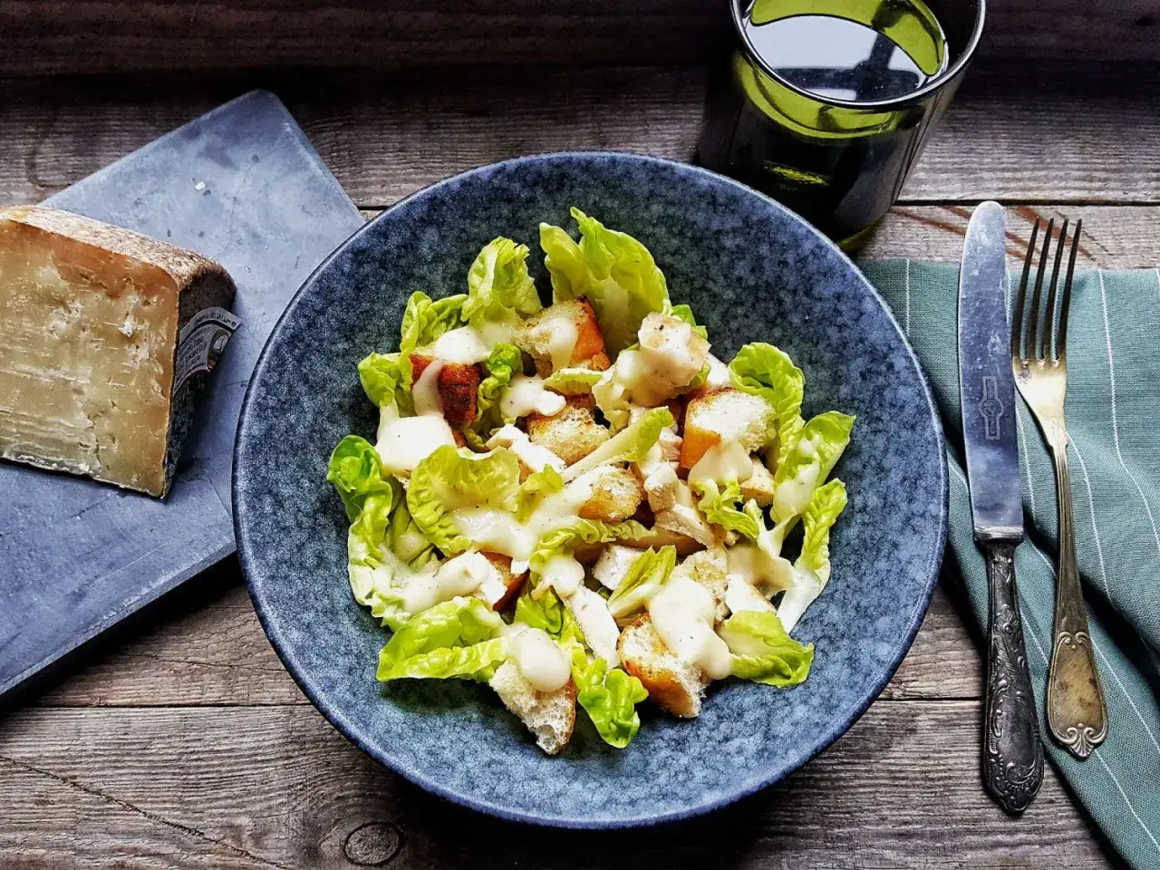 Insalata Caesar con crostini e una generosa spolverata di crema di pecorino, accanto a un pezzo di formaggio stagionato.