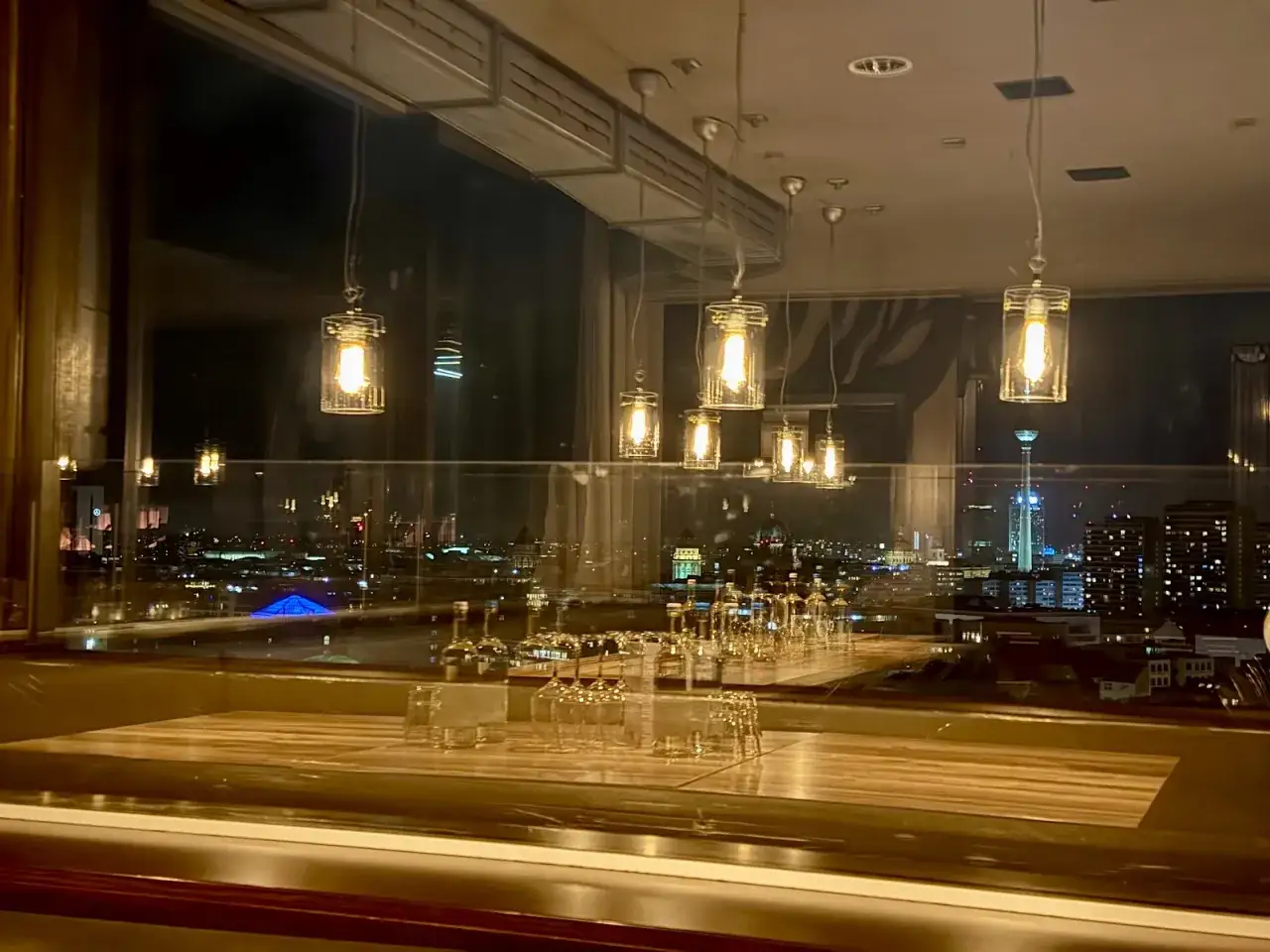 Stimmungsvoller Blick auf die Skyline Berlins bei Nacht, mit Gl&auml;sern und Lampen im Vordergrund. Ein perfekter Ort f&uuml;r die besten Restaurants in Berlin.