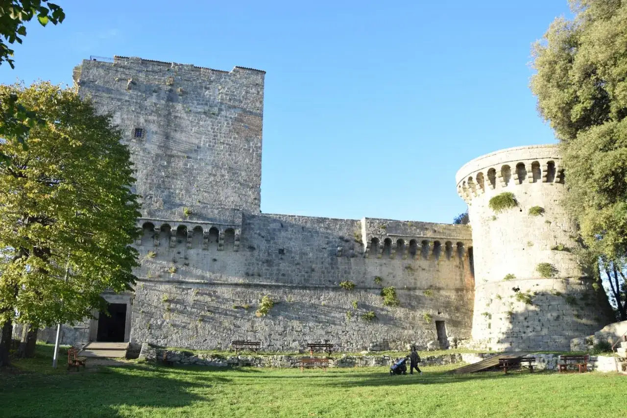 Il castello di Sarteano, con la sua imponente torre e mura antiche, si staglia contro il cielo azzurro. Un prato verde invita alla sosta.