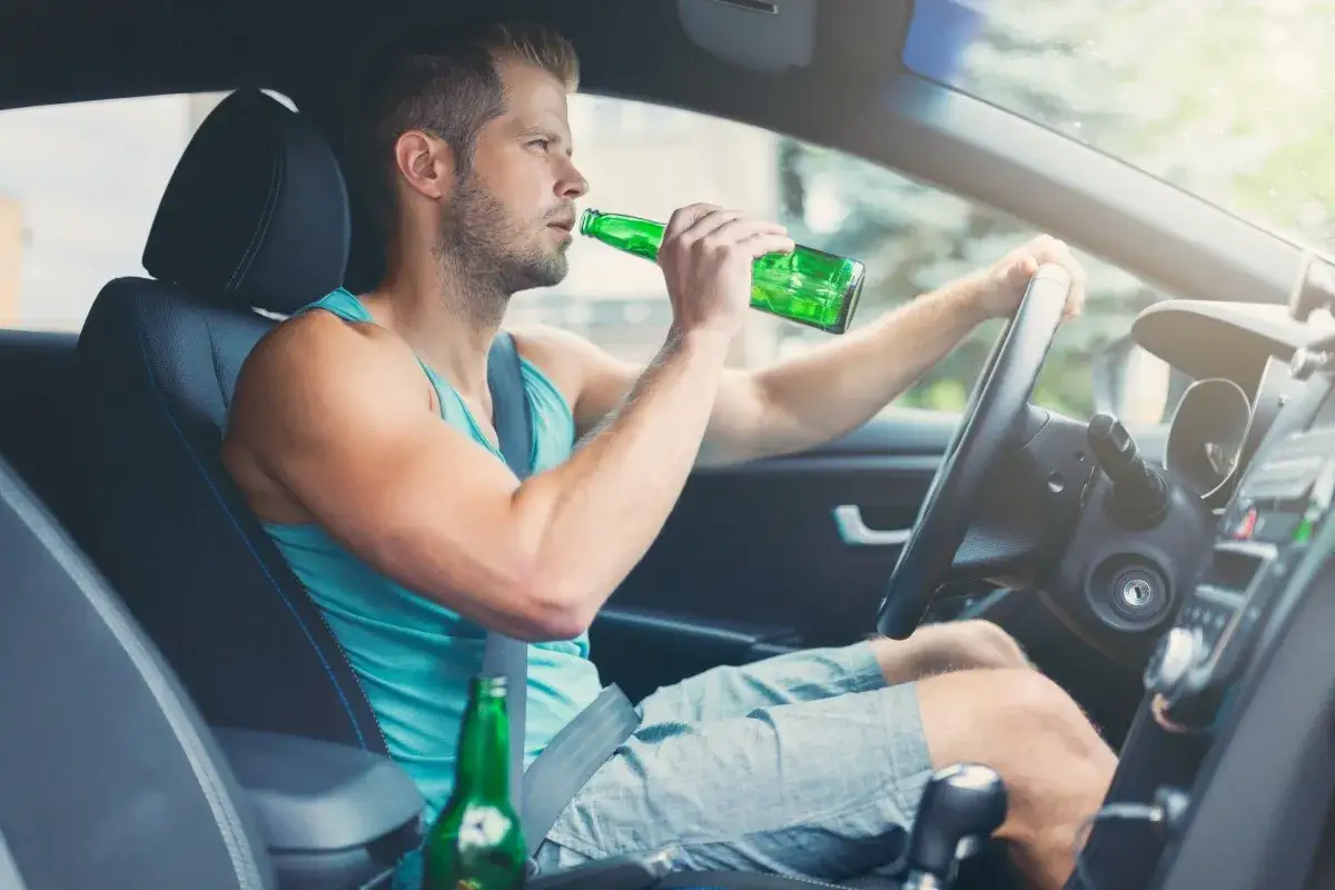 Mężczyzna pije piwo za kierownicą. Niebezpieczna jazda, która może wymagać analizy czasu trzeźwienia po alkoholu, tabela.