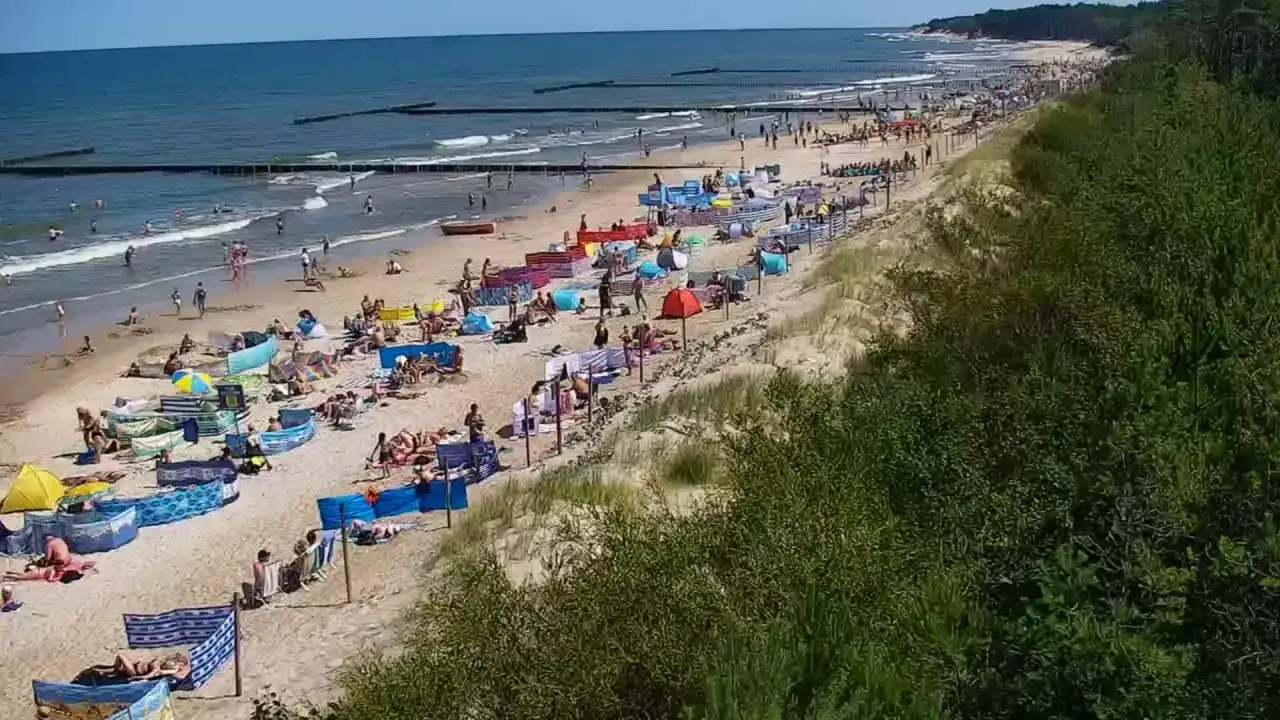 Słoneczna plaża w Jarosławcu tętni życiem. Ludzie odpoczywają na piasku, kąpią się w morzu i spacerują wzdłuż brzegu. W oddali widać falochrony.