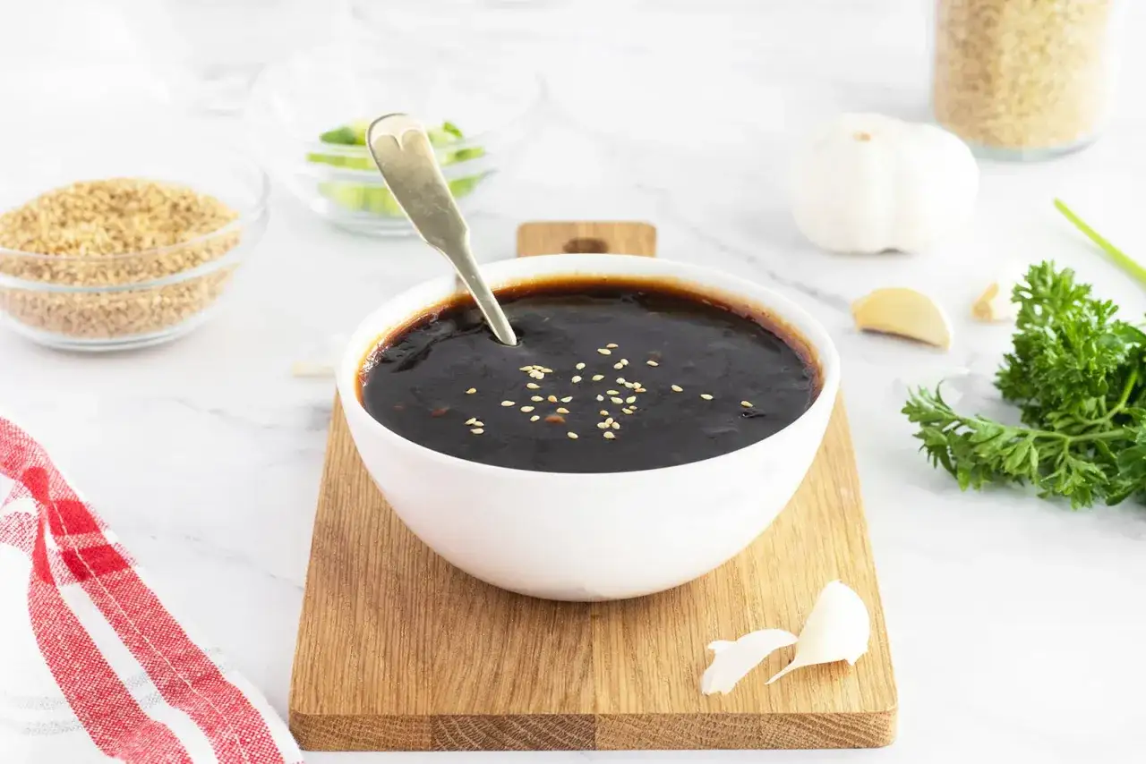 Sos teriyaki przepis: ciemny, błyszczący sos w białej misce, posypany sezamem. Obok składniki: czosnek, natka pietruszki, sezam.