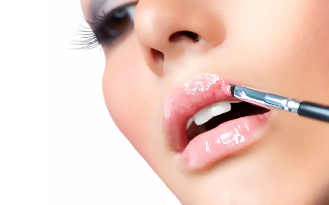 Dettaglio di labbra lucide e rosa, con un pennello che applica gloss. Un'immagine che mostra come mettere il rossetto per un effetto voluminoso.