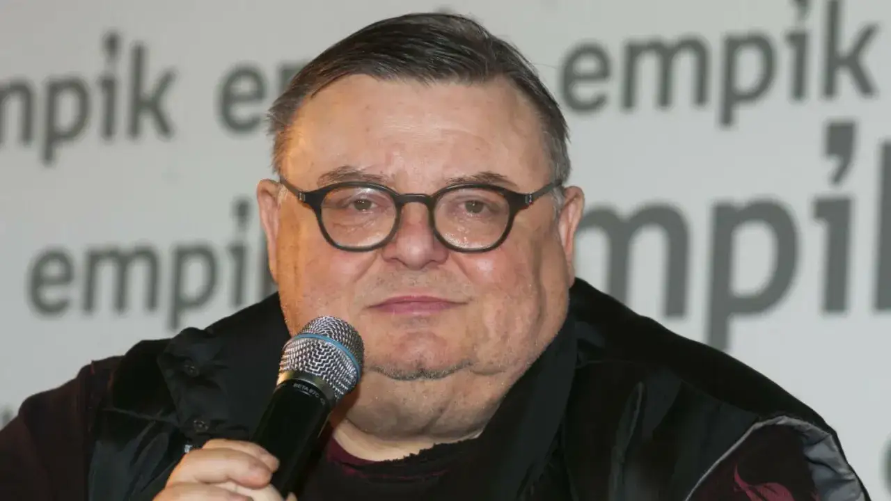 Wojciech Mann w okularach, z mikrofonem w dłoni, na tle napisu