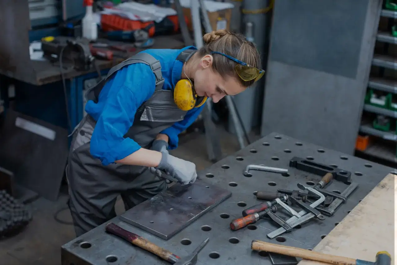 Mujer con equipo de protecci&oacute;n trabajando en un **taller ordenado**. Herramientas y piezas met&aacute;licas sobre la mesa de trabajo.