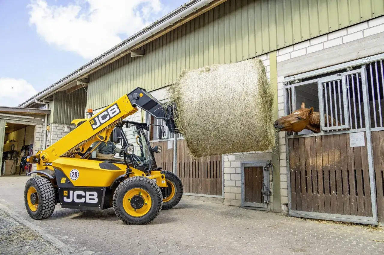 Żółta ładowarka teleskopowa JCB podnosi belę siana do stajni, gdzie koń wygląda z okna.