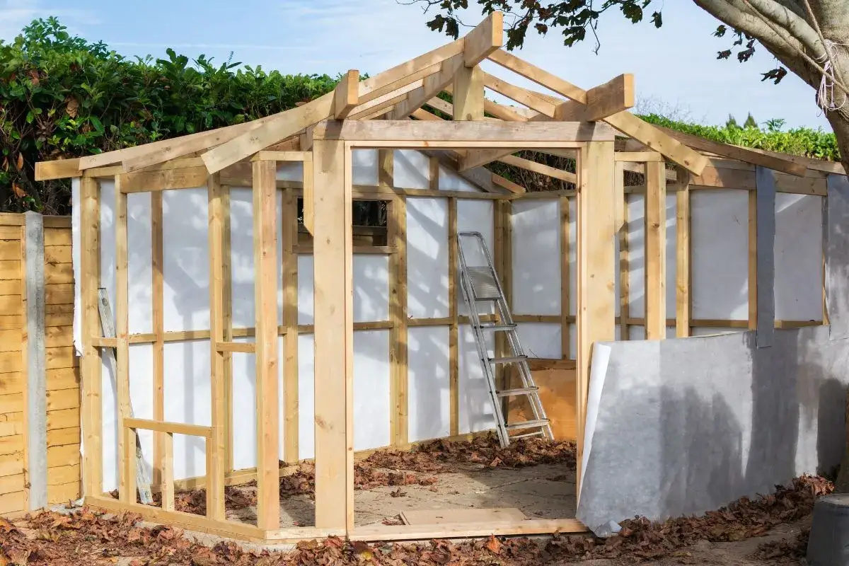 Structure en bois d'une cabane de jardin en construction, avec un toit en cours d'installation et une échelle à l'intérieur.