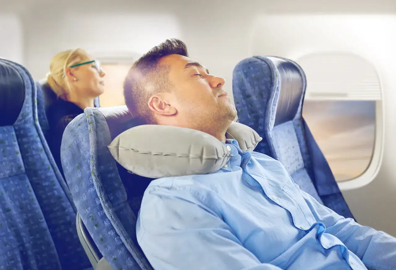 Mann schl&auml;ft im Flugzeug mit Nackenkissen, um Jetlag zu vermeiden. Eine Frau sitzt daneben.