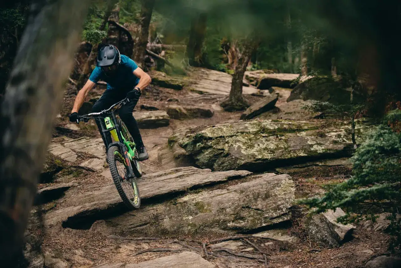 Troy Brosnan navigates a rocky, wooded trail on his mountain bike, expertly maneuvering over obstacles.