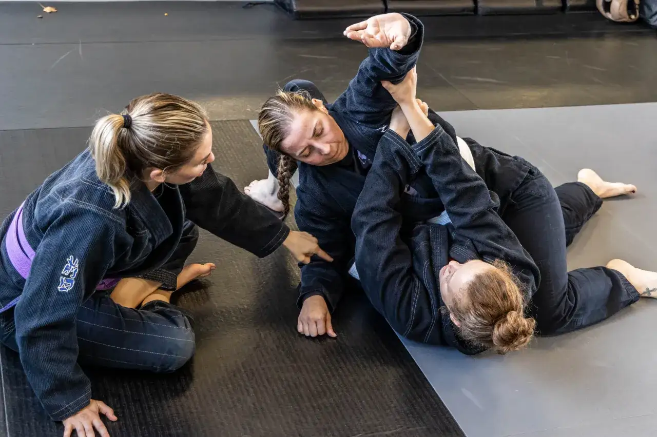 Tre kvinnor i gi tränar combat jiu jitsu på en matta. En kvinna håller armen på en annan som ligger ner.