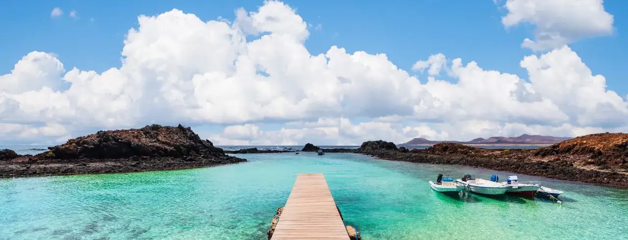 Muelle de madera hacia aguas turquesas y barcas. Un para&iacute;so para descubrir qu&eacute; ver en Fuerteventura.
