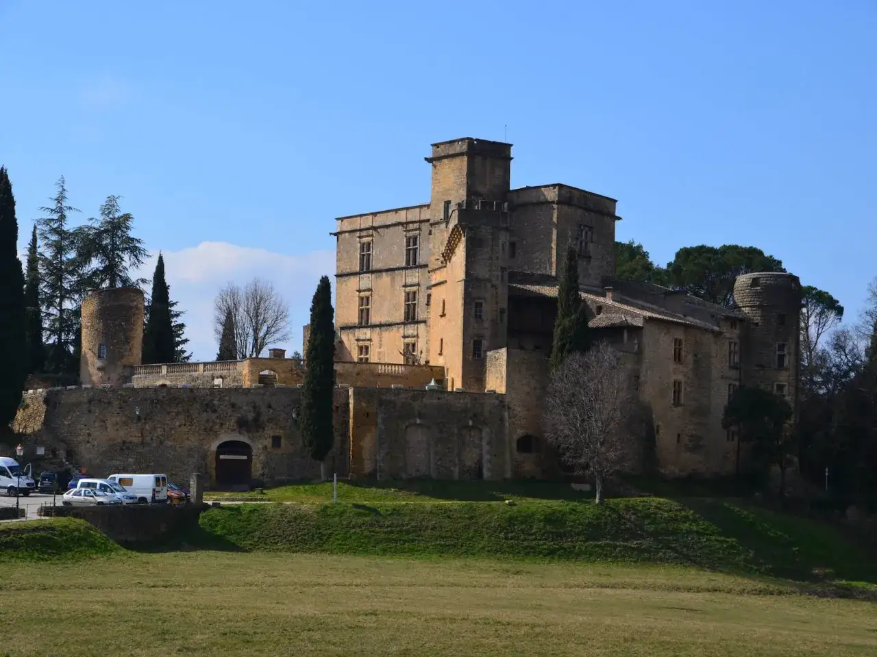 Le majestueux **Ch&acirc;teau de Lourmarin** se dresse sur une colline, entour&eacute; de cypr&egrave;s et d'arbres. Un ciel bleu clair domine le paysage.
