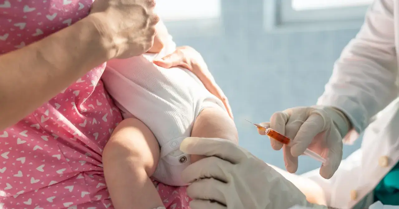 Pierwsze szczepienie niemowlaka. Lekarz w rękawiczkach podaje zastrzyk. Mama tuli dziecko.