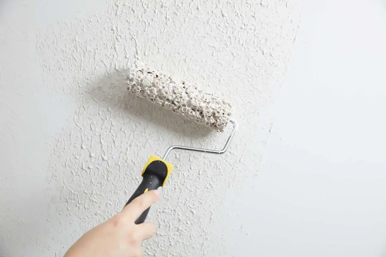 En hand håller i ett handtag till en roller som används för att applicera rullspackel på en vägg, vilket skapar en texturerad yta.