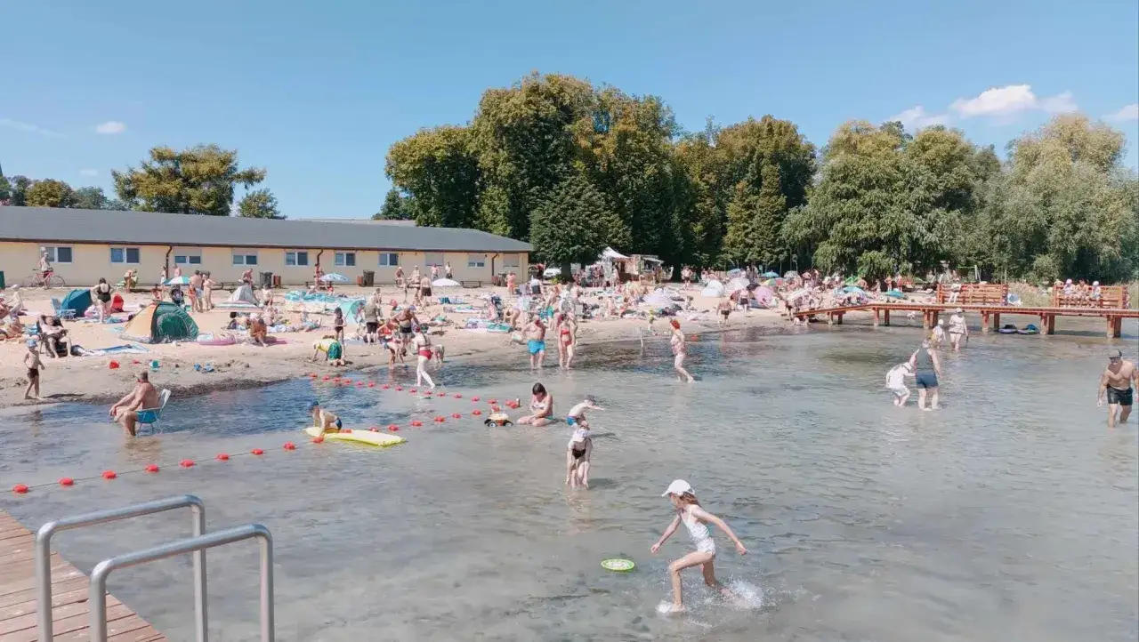 Tłum ludzi odpoczywa nad wodą, dzieci bawią się na plaży i w płytkiej wodzie. Jezioro Powidzkie tętni życiem w słoneczny dzień.