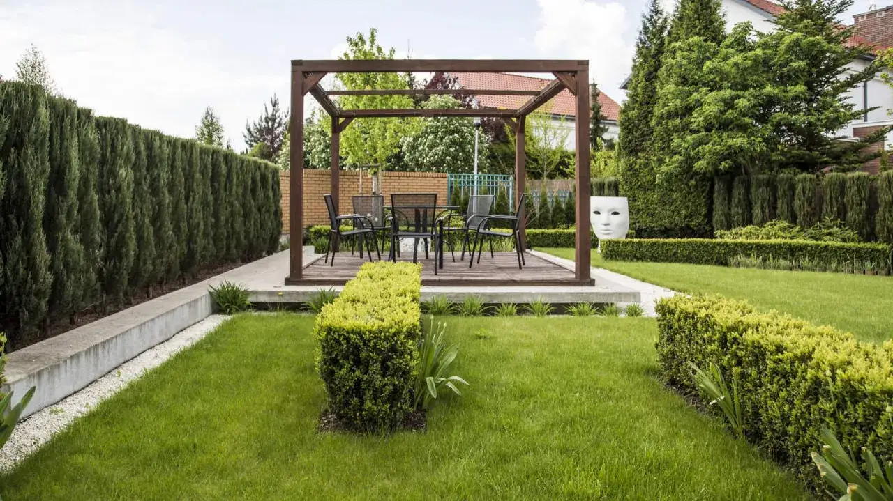 Nowoczesne aranżacje ogrodu: drewniana pergola z meblami, wysokie żywopłoty i rzeźba maski tworzą stylową przestrzeń.
