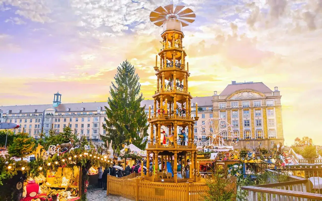 Der **beste Weihnachtsmarkt Deutschlands** mit einer riesigen Holzpyramide, geschmücktem Baum und Buden unter einem farbenfrohen Himmel.
