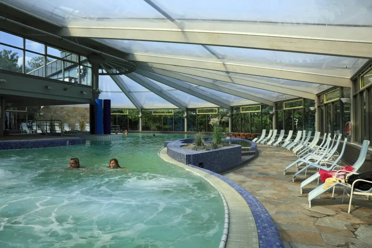Zwei Frauen schwimmen im **Mattlerbusch Schwimmbad** unter einem transparenten Dach. Liegestühle laden zum Entspannen ein.