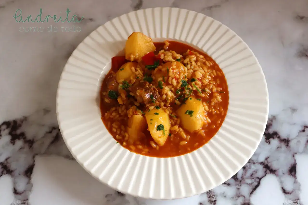 Un plato de arroz con trozos de costillas y patatas, todo ba&ntilde;ado en una salsa rojiza y espolvoreado con perejil picado.