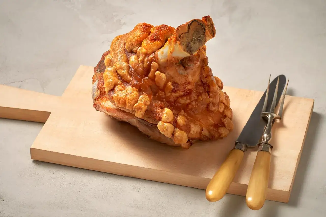 Knusprige Schweinshaxe auf Holzbrett, bereit für ein köstliches Schweinshaxe Rezept. Messer und Gabel liegen daneben.