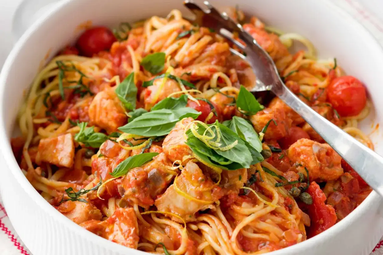 Makaron z rybą w sosie pomidorowym, ozdobiony bazylią i skórką cytrynową. Widać też zielone **kapary do czego** dodają daniu wyrazistości.
