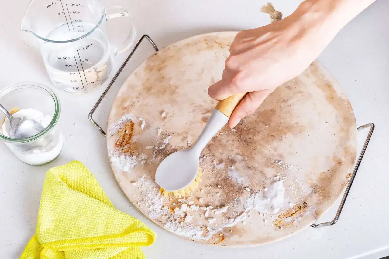 Czyszczenie kamienia do pizzy sodą oczyszczoną i wodą. Dłoń szoruje kamień szczotką, obok stoi dzbanek z wodą i słoik z sodą.