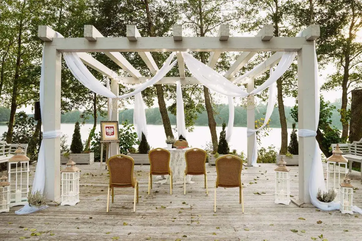 Ustawienie na ślub cywilny na luzie nad jeziorem. Biała pergola, krzesła, ozdoby i polski herb tworzą romantyczną atmosferę.